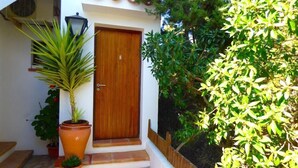 A wooden door with a small porch and steps leading up to it. There are potted plants on the porch and a wall-mounted air conditioning unit to the left. - Sa Pergola (Formentera)