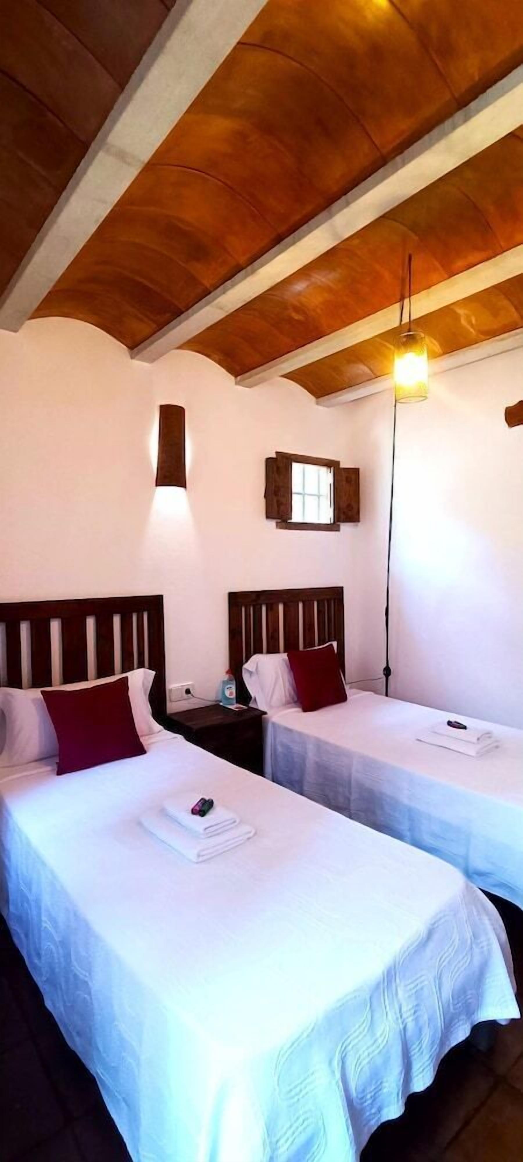 A room with two beds, wooden headboards, a small window, and a hanging light fixture.
