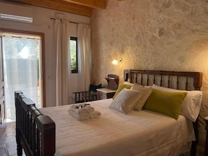 Un dormitorio con cabecera de madera, cama con almohadas blancas y verdes, mesita de noche con lámpara y vista a un jardín por una ventana.