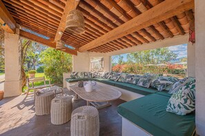 Area relax esterna coperta, con soffitto in legno, mobili in vimini e vista sul verde.