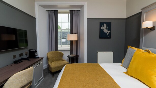 Habitación de hotel con cama, escritorio, silla, televisor, lámpara y ventana con cortinas.