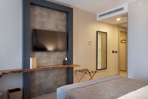 Een moderne hotelkamer met een flatscreen televisie bevestigd aan een gestructureerde muur, een houten plankje met een zakdoos, een hangende handdoekhouder, en een deur met een nummerbord.