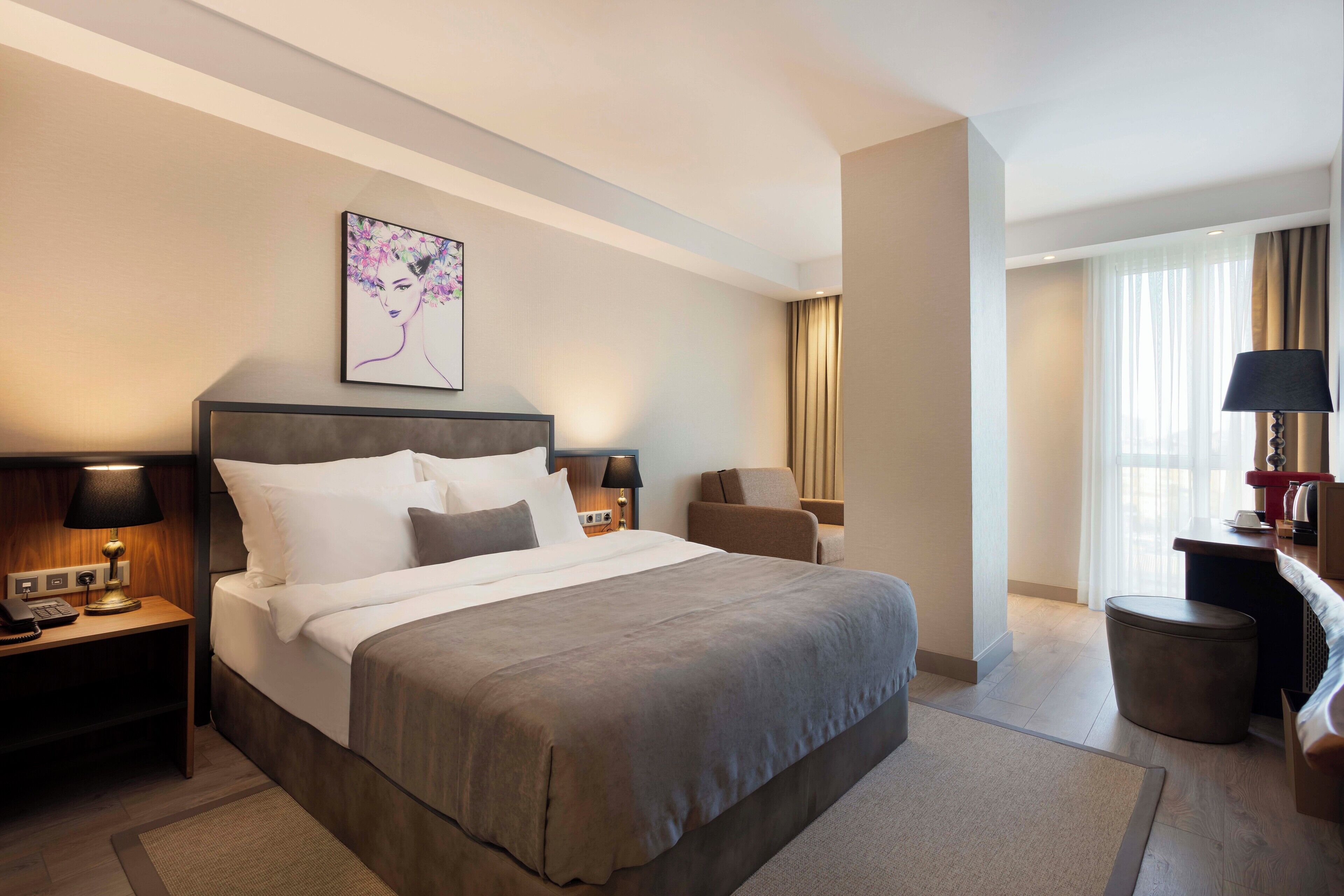 Ein Hotelzimmer mit einem großen Bett, einem Schreibtisch inklusive Lampe, einem Sessel, einem Nachttisch und einem großen Fenster mit Vorhängen.