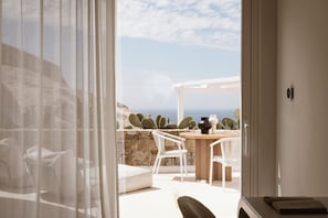 Una habitació amb vistes al mar, una cadira blanca, una taula amb un vas i una planta de cactus.