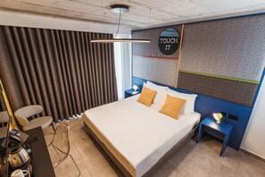 Ein modernes Hotelzimmer mit einem großen Bett, einem Schreibtisch mit Kaffeemaschine, einem Sessel und einem an der Wand montierten Fernseher.
