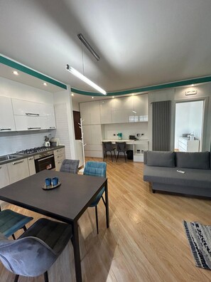 Una cocina moderna con armarios blancos, una zona de comedor con una mesa oscura y sillas azules, y un sofá gris en la sala de estar.