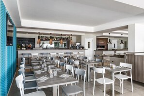 A modern restaurant with multiple dining tables and chairs, a bar area with bottles, and a kitchen visible in the background. - Hotel Centosedici (Terracina)