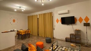 A living room with a sofa, ottoman, dining table, chairs, curtains, a TV, and a patterned rug. - Hotel Centosedici (Terracina)