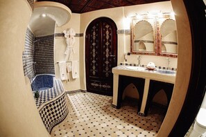 A bathroom with a checkered floor, a bathtub, a sink with a mirror, and a dark wooden door. - OZ Palace Ouarzazate (Tarmigt)
