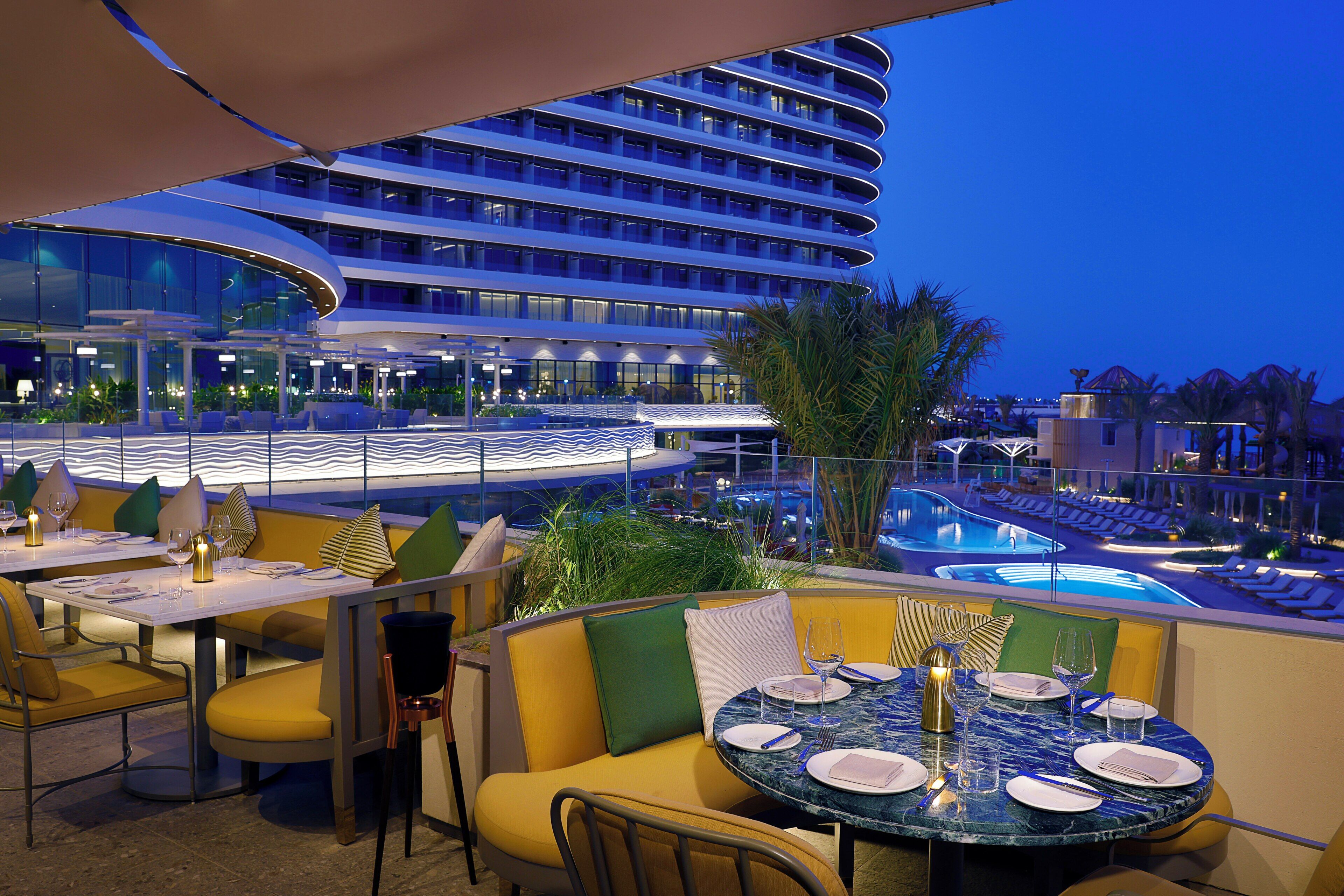 A rooftop dining area with tables set for two, overlooking a pool and a modern building.