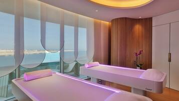 A modern spa room with two treatment beds, large windows, and a view of the sea.