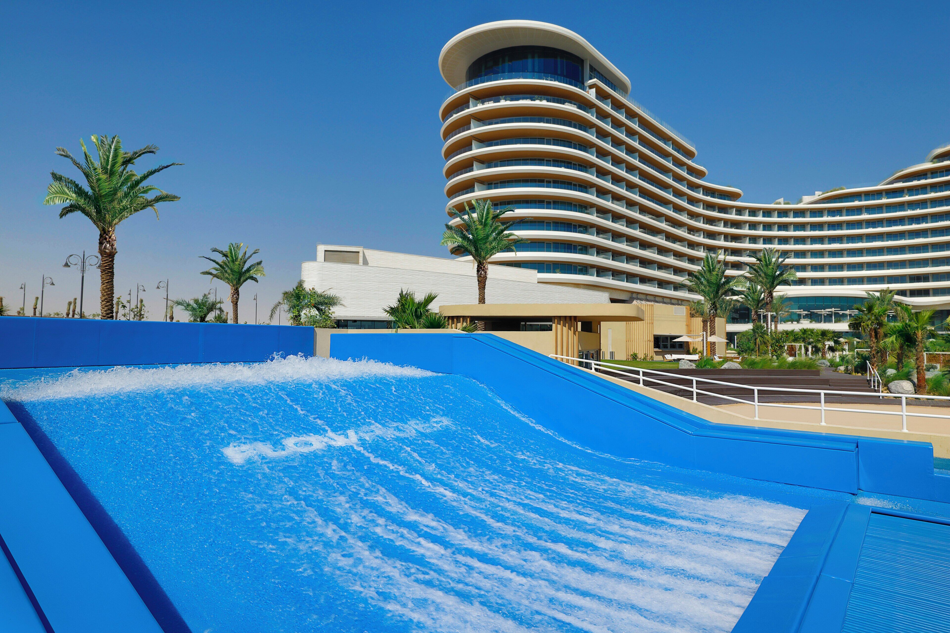 Un hotel con arquitectura curvilínea, una piscina con efecto de olas, palmeras y un cielo azul despejado.
