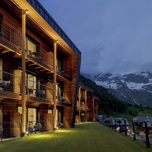 Un bâtiment en bois à plusieurs étages, doté de balcons, bordé d’un sentier herbeux et ayant une chaîne de montagnes en toile de fond.