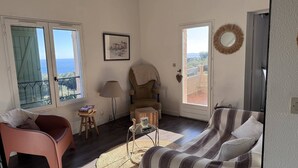 Una sala de estar con vistas al mar, una silla, un sofá, una lámpara y una mesita con libros.