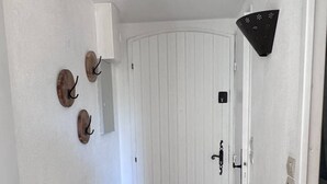 Un pasillo con una puerta blanca, un aplique de pared y un estante metálico con cestas y objetos decorativos.
