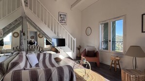 Un salón espacioso con una cama grande, una escalera que sube a un piso superior, un televisor y vistas al exterior a través de un ventanal amplio.