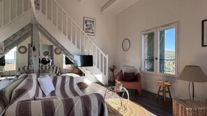 Ein geräumiges Wohnzimmer mit einem großen Bett, einer Treppe, die zu einer oberen Etage führt, einem Fernseher und Blick ins Freie durch ein großes Fenster.