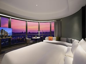 A modern hotel room with a curved ceiling, a large bed, a sofa, a small table, and a TV displaying a cityscape. - Novotel Shanghai Wanyuan (Shanghai)