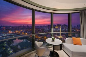 A modern room with a curved glass wall offering a city view at dusk. - Novotel Shanghai Wanyuan (Shanghai)