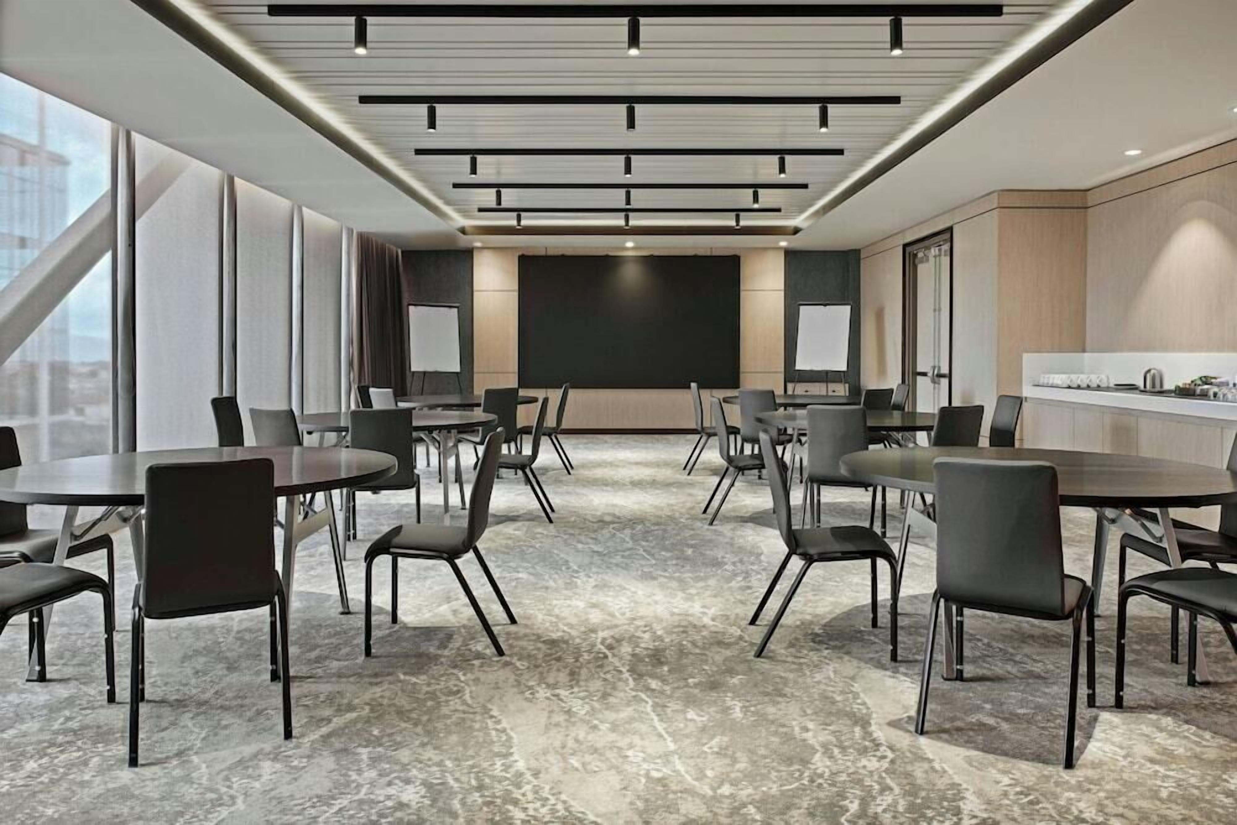 Una sala de conferencias moderna con mesas y sillas redondas, una pantalla grande y una zona de cocina.