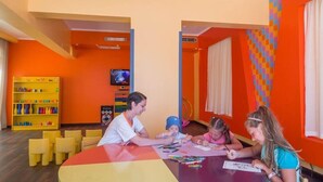 A brightly colored room with orange walls, a table with children and adults engaged in activities, and a yellow bookshelf filled with toys. - Sunny Days Palma De Mirette Resort (Hurghada)