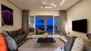 A modern living room with a sofa, a coffee table, a flat-screen TV, and a view of the outdoors through large windows. - The Mora Zanzibar (Matemwe)
