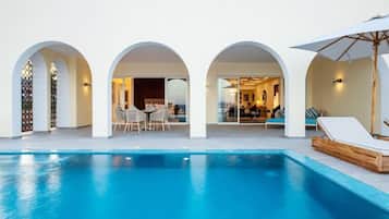 A swimming pool with arched walkways, a dining area, and a lounge with a sun umbrella.