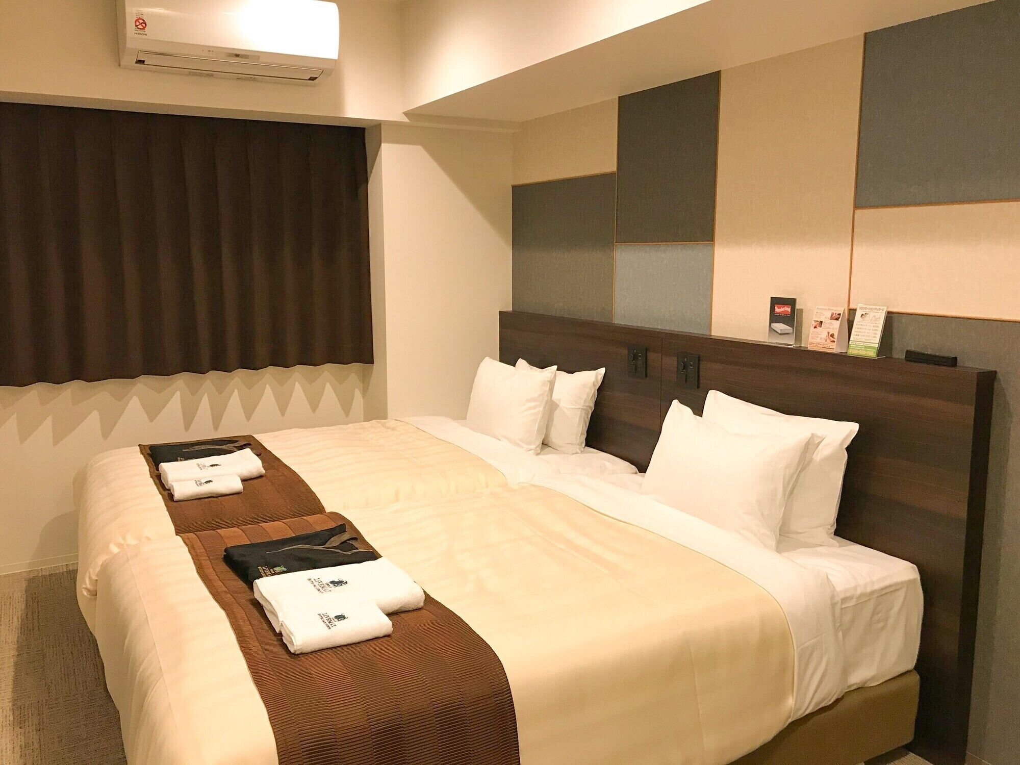 Ein Hotelzimmer mit einem großen Bett, einem Kopfteil mit Stauraum, einem Fenster mit Vorhängen und einer Klimaanlage.