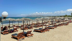 A beach with rows of wooden sun loungers and thatched umbrellas, a clear blue sky, and the sea in the background. - Mare Blue and Suites (Apokoronas)