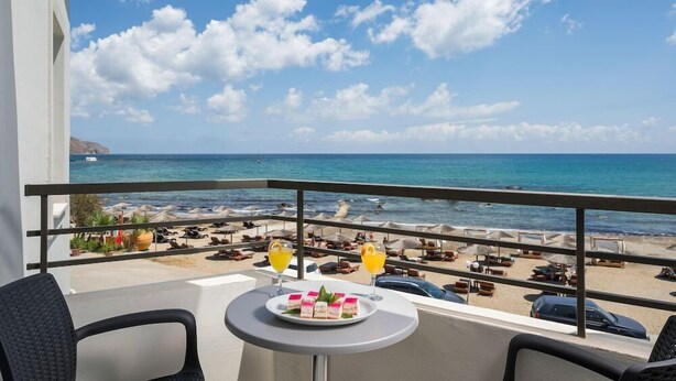 A balcony with a table set for two, overlooking a beach with umbrellas and lounge chairs. - Mare Blue and Suites (Apokoronas)