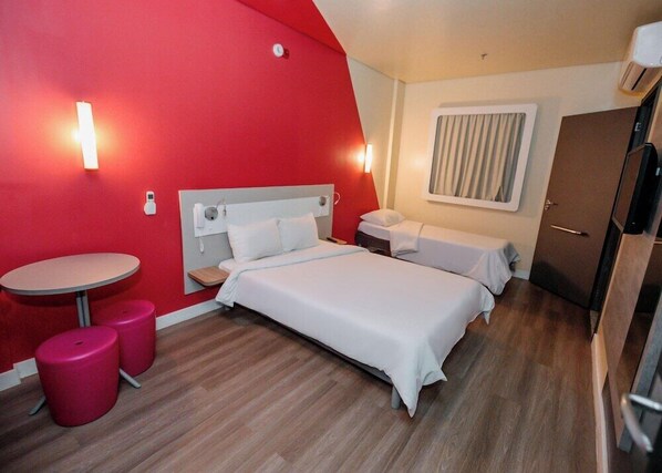 Une chambre d’hôtel avec un mur d’accent rouge, un lit avec une literie blanche, une petite table ronde avec deux tabourets roses, une table de chevet avec une lampe et une fenêtre avec des rideaux.