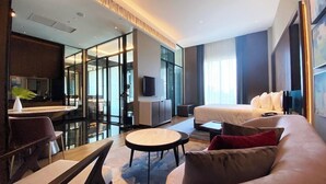 Une chambre d’hôtel moderne dotée d’un grand lit, d’un coin salon avec une table ronde, d’une télévision et d’une paroi en verre séparant l’espace de vie de la salle de bain.