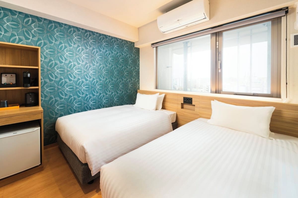 A hotel room with two beds, a minibar, a desk with a coffee maker, a window with a view, and a patterned wall.