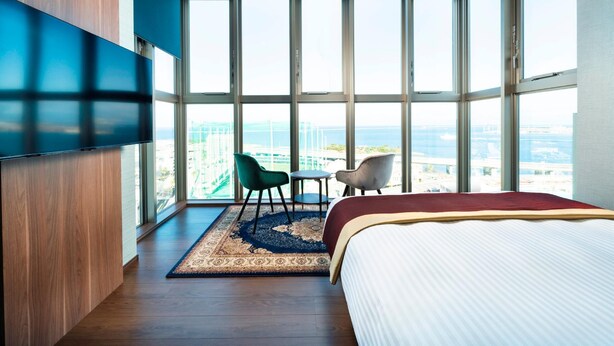 Une chambre d’hôtel avec une grande fenêtre, un lit, une télévision et un coin salon avec des fauteuils.