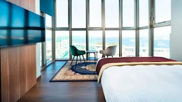 A hotel room with a large window, a bed, a TV, and a seating area with chairs.