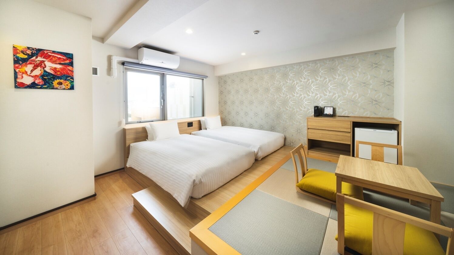 A compact hotel room with two beds, a desk, a chair, a small table, and a window.