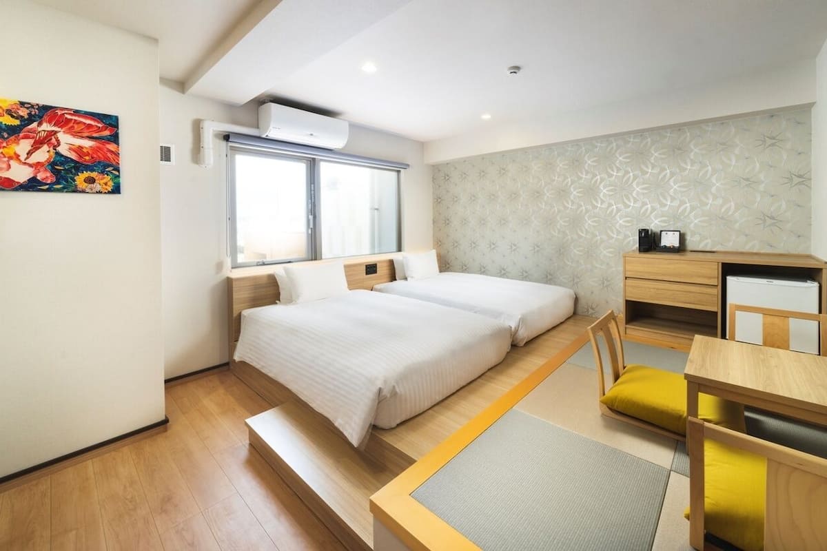 A compact hotel room with two beds, a desk, a chair, a small table, and a window.