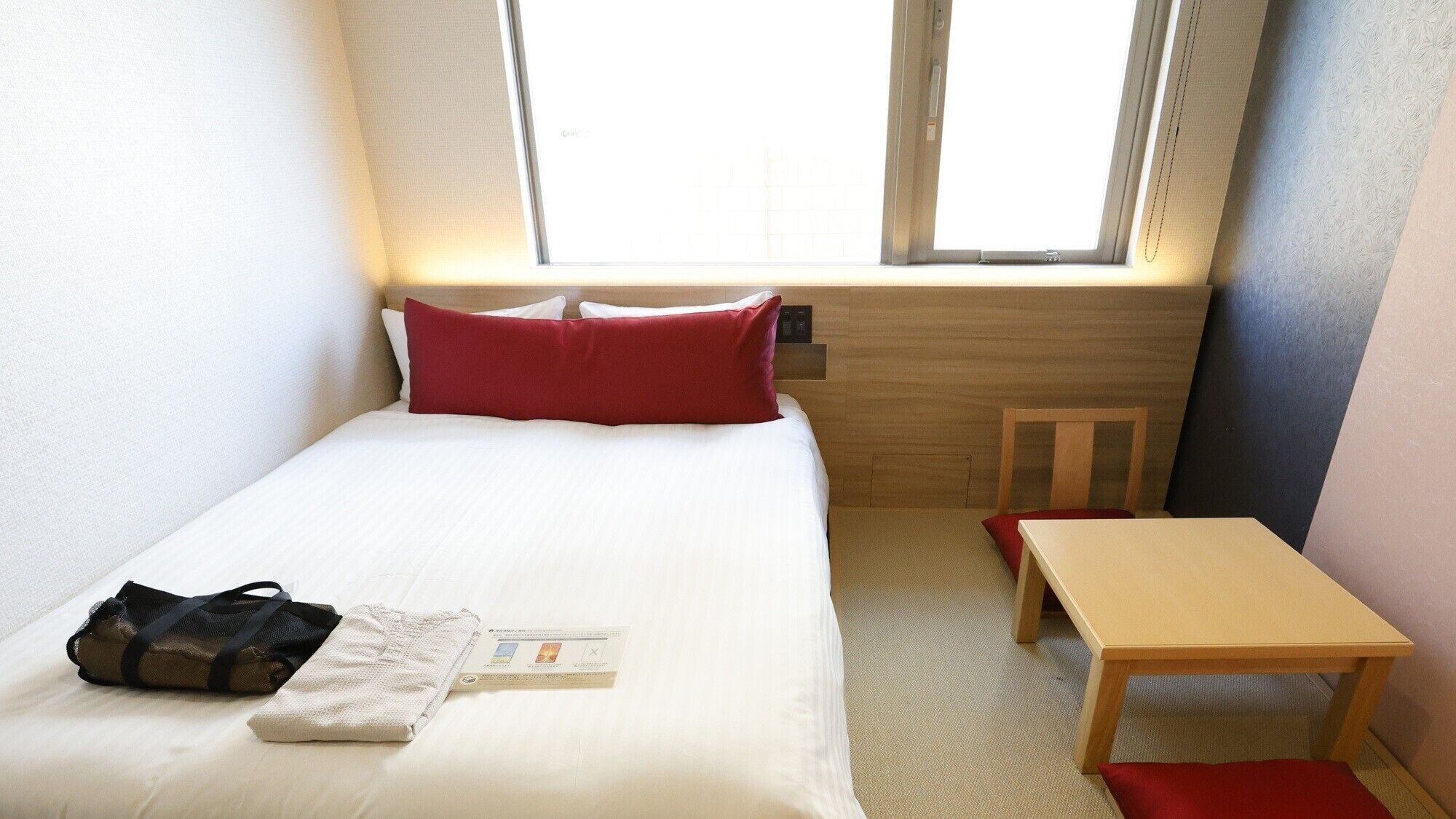 A small, neatly arranged hotel room with a bed, a bedside table, a chair, and a window.