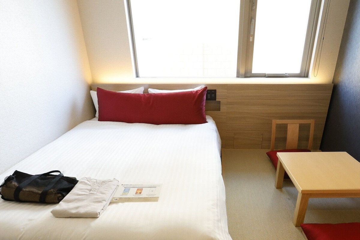 A small, neatly arranged hotel room with a bed, a bedside table, a chair, and a window.