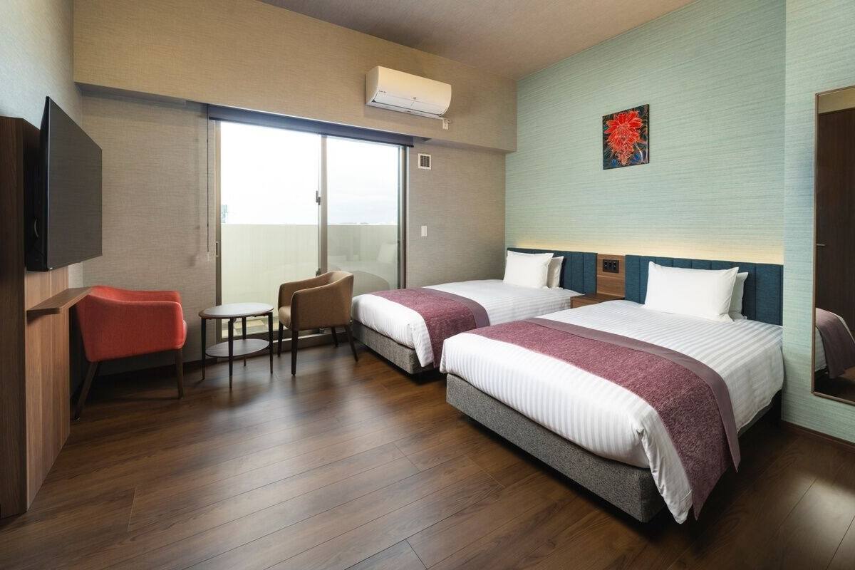 A hotel room with two beds, a TV, a chair, a small table, and a sliding door to a balcony.