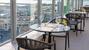 Un restaurant situé dans un immeuble de grande hauteur, offrant une vue panoramique sur la ville et la mer, avec des tables rondes et des chaises en osier.