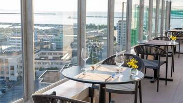 A high-rise restaurant with panoramic city and sea views, featuring round tables and wicker chairs.