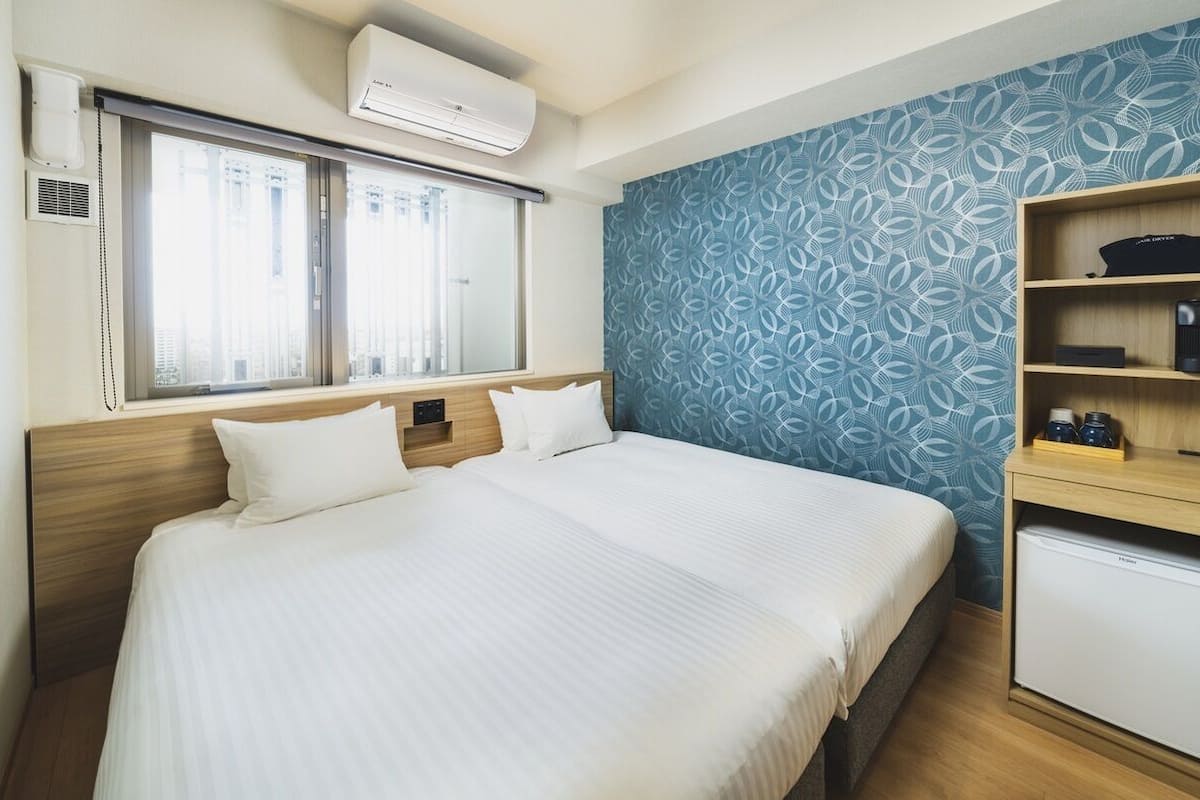 A hotel room with a bed, a desk with a refrigerator, and a window with blinds.