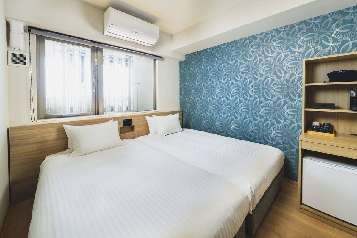 Hotellihuone, jossa on sänky, työpöytä, jossa on jääkaappi, ja ikkuna, jossa on sälekaihtimet.