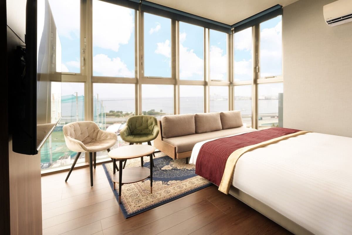 A hotel room with a large window, a bed, a sofa, a chair, and a small table.