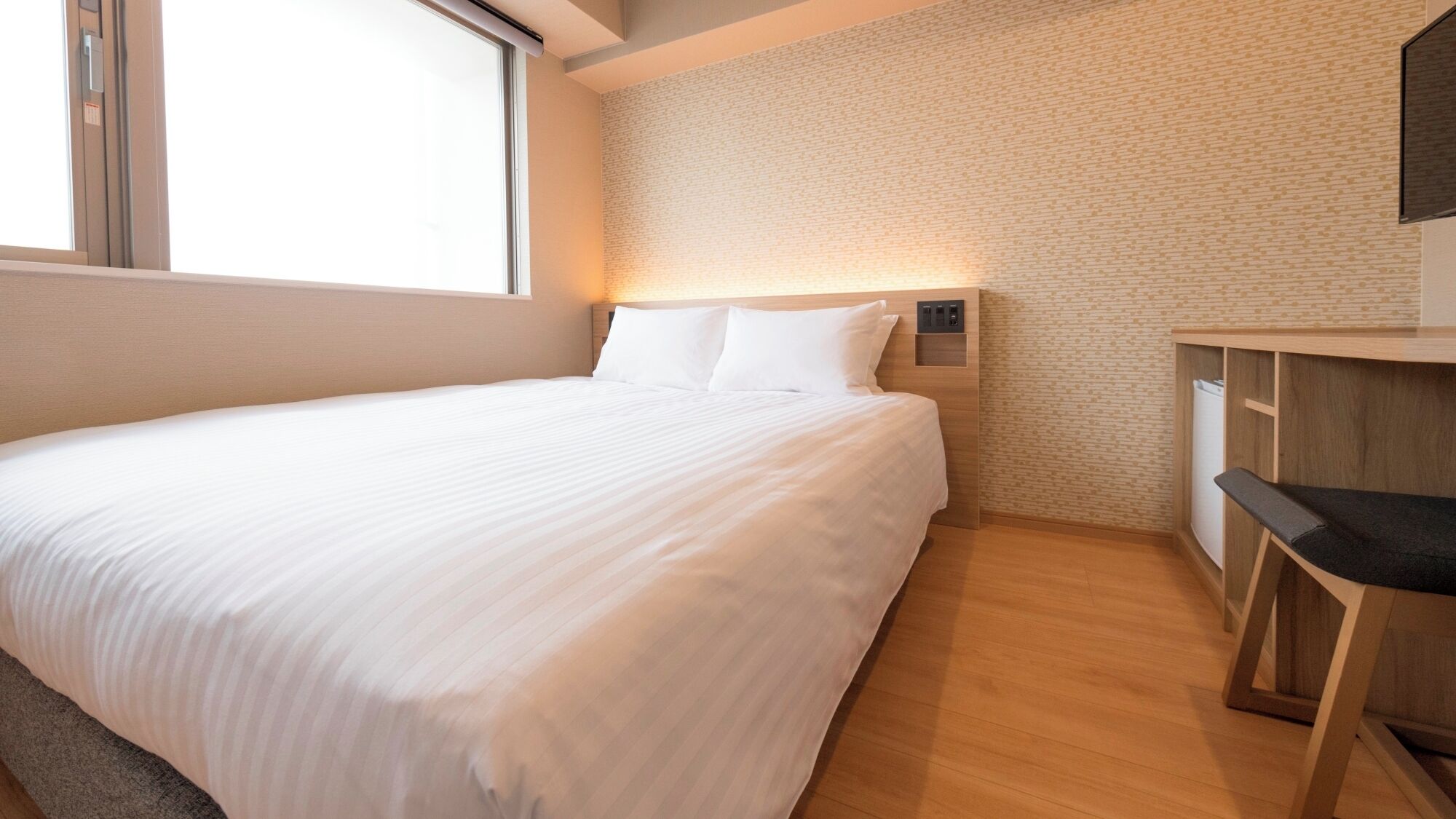 A neatly made bed in a room with a brick-patterned wall, a small desk with a chair, and a window providing natural light.