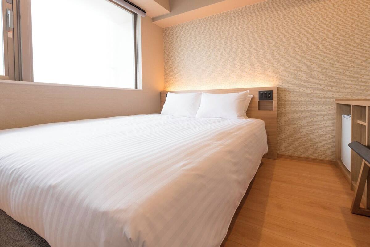 A neatly made bed in a room with a brick-patterned wall, a small desk with a chair, and a window providing natural light.