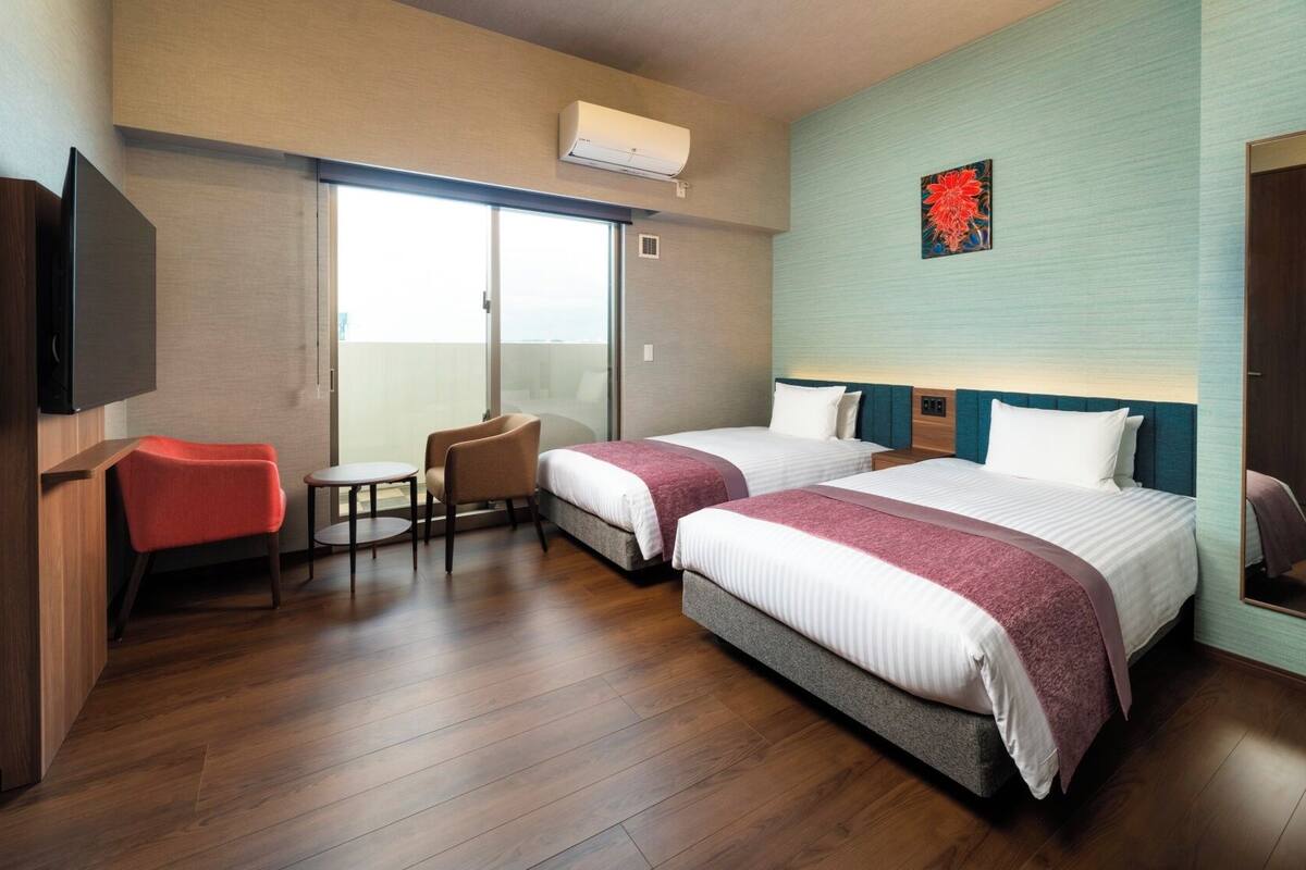 A hotel room with two beds, a TV, a chair, a table, and a balcony.