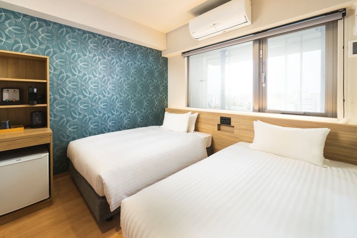 A hotel room with two beds, a small kitchenette, a built-in TV, and a large window.