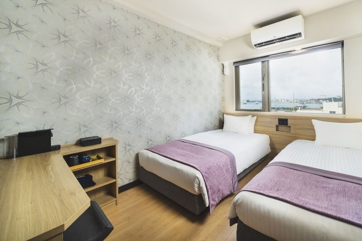 A hotel room with two beds, a desk, a shelf with items, and a large window with a view.
